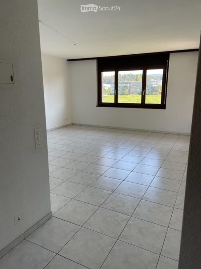 Empty room with tiled floor, white walls, and a large window with a view of greenery outside.