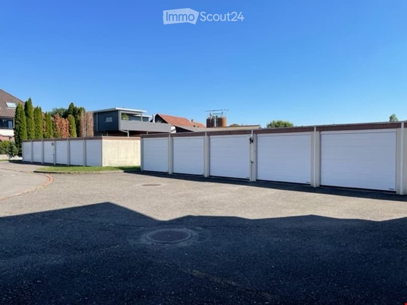 A row of white garage units with a modern design, situated in a residential area with trees and neighboring houses visible in the background.