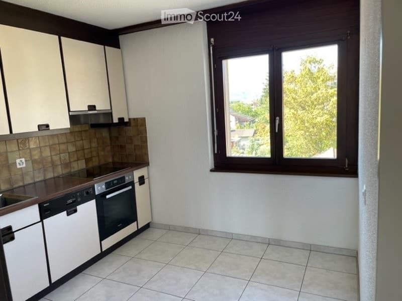 Modern kitchen with white cabinets, tiled backsplash, and a large window offering a view of greenery outside. Includes a sink, dishwasher, and oven.