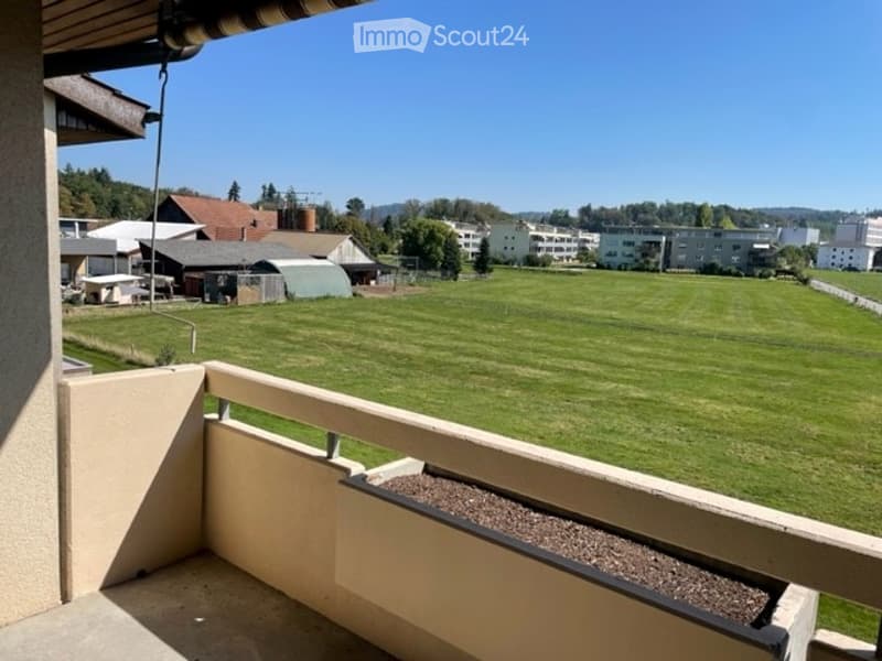 Balcony overlooking a large green field with distant buildings and trees under a clear blue sky.