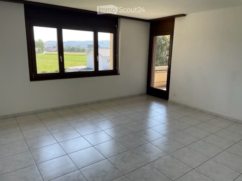Empty room with tiled floor, large window overlooking a green field, and a sliding door leading to a balcony.