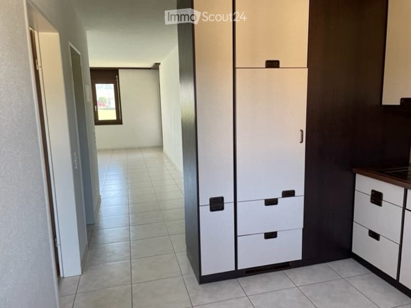 A clean, modern hallway with tiled flooring, white walls, and built-in storage units. A window at the end provides natural light.