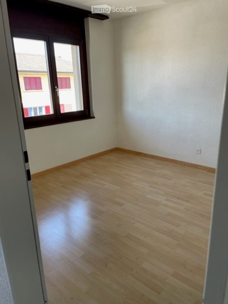 Empty room with wooden flooring and white walls, featuring a large window with brown frame and a view of a neighboring building.