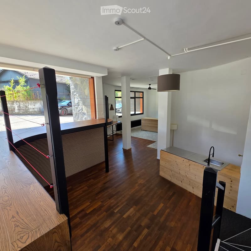 Modern, minimalist room with wooden flooring, white walls, and a sink area. Features include a hanging lamp, a small table with a chair, and a stool near the window.