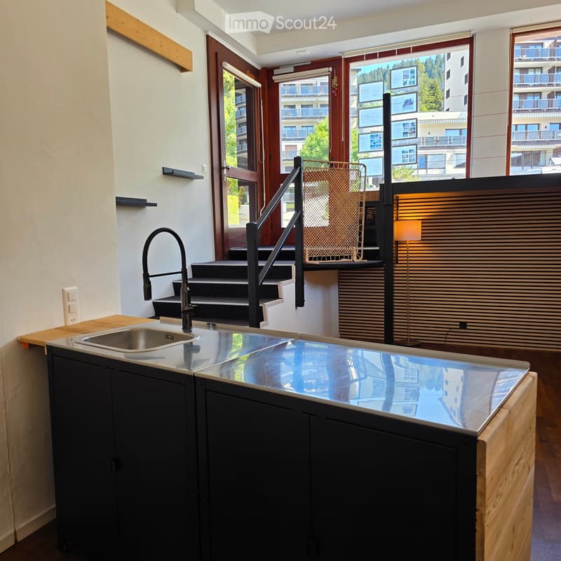 Modern kitchen with a sleek black cabinet, stainless steel sink, and a reflective countertop. Features a staircase leading upwards and large windows offering a view of the building exterior.