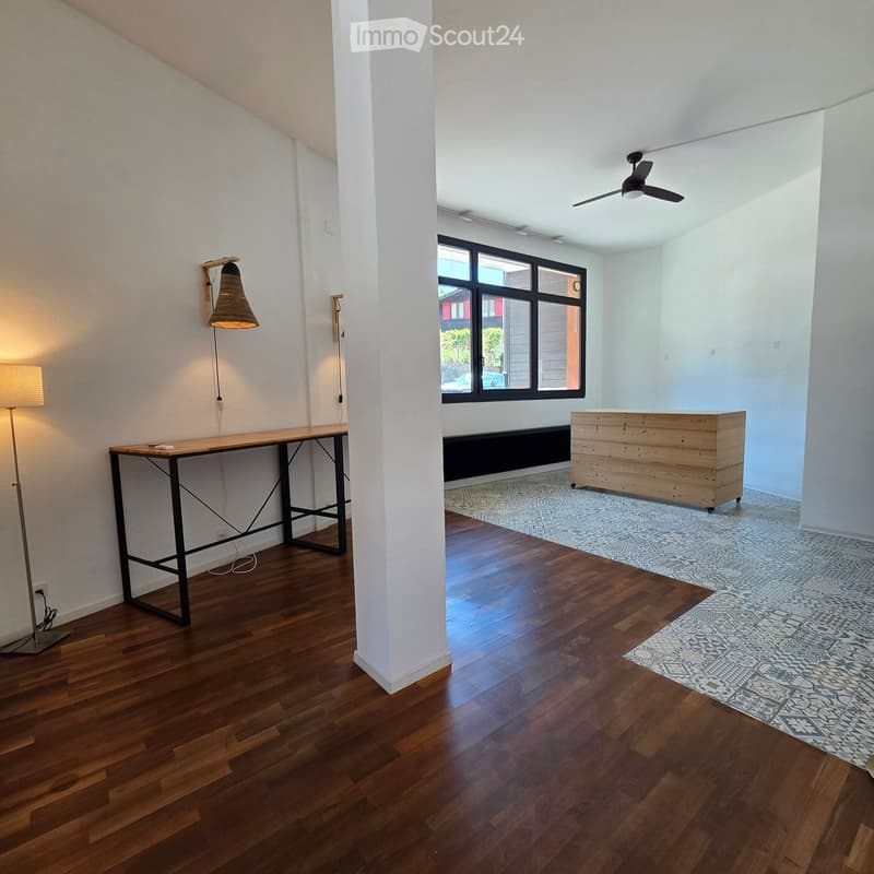 A spacious, well-lit room with wooden flooring, a large window, a ceiling fan, a long wooden table with a black metal base, a tall floor lamp, a wall-mounted lamp, and a wooden cabinet.