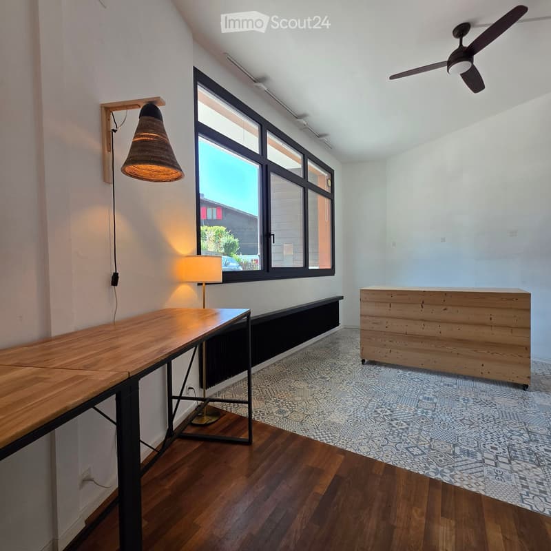 Bright, modern room with wooden flooring, large windows, a long wooden table, a lamp, and a wooden cabinet.