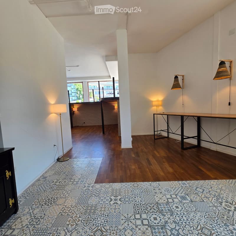 Modern, minimalist room with wooden flooring, white walls, and geometric patterned tiles. Features a long black-framed table, floor lamp, and wall-mounted lamps.