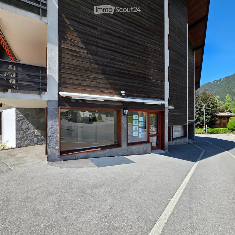 Modern building with wooden facade, stone base, and glass entrance. Features a balcony on the left and a clear view of the surrounding landscape.