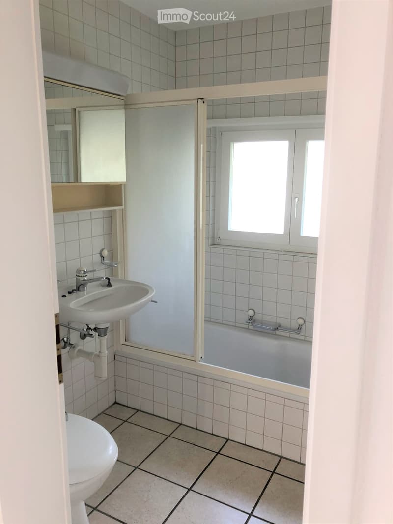 Bathroom with a sink, toilet, bathtub, mirror, and cabinet. The walls are tiled, and there is a window above the bathtub.