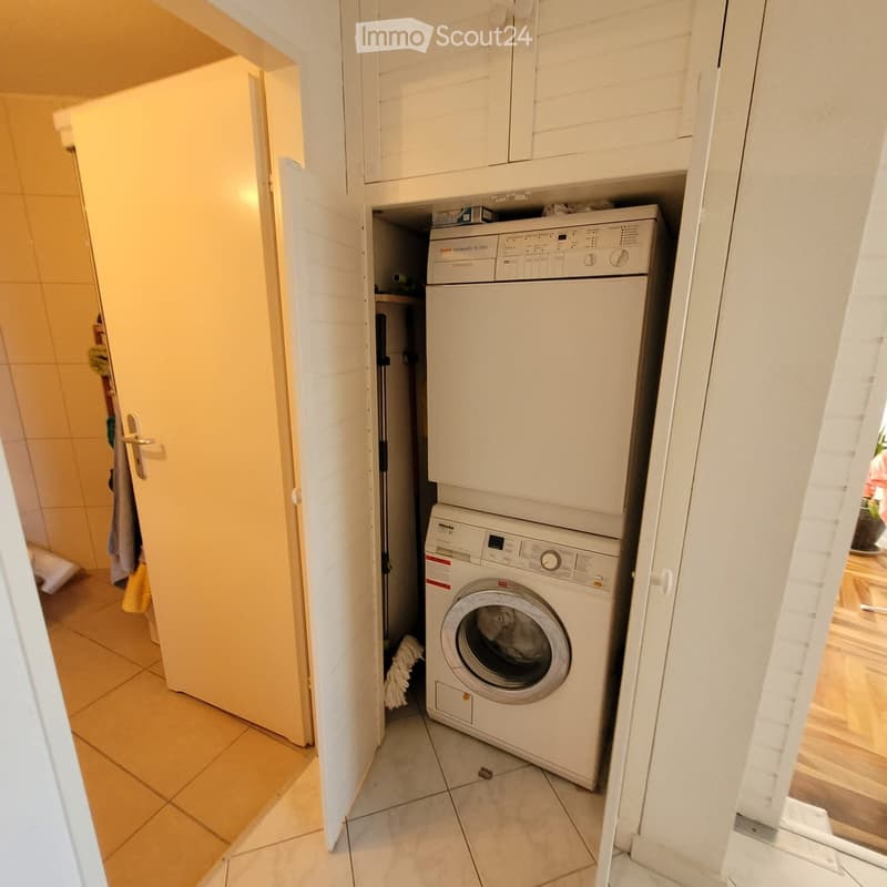 Small bathroom with a white washing machine and dryer stacked vertically, tiled floor, and white cabinets.