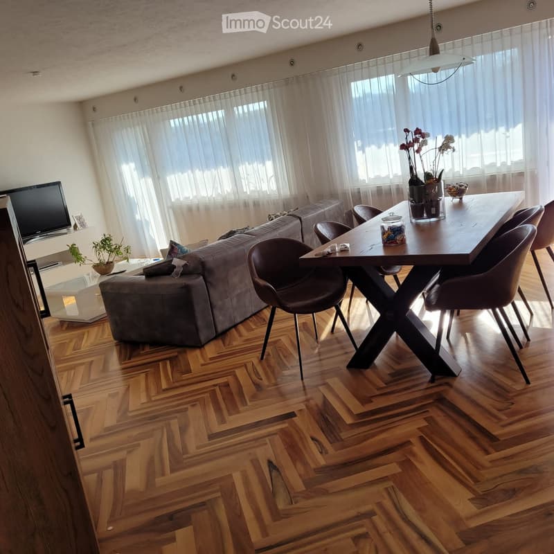 Modern living and dining area with wooden herringbone flooring, large windows with sheer curtains, grey sectional sofa, glass coffee table, dark wood dining table with chairs, and decorative plants.