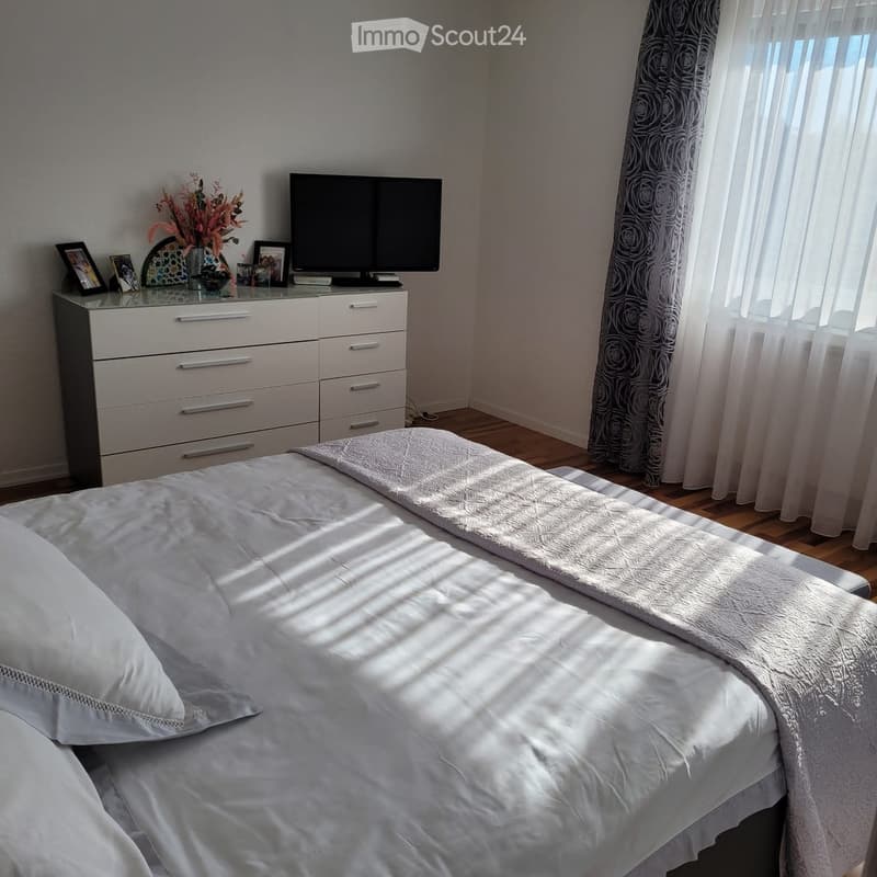 A well-lit bedroom with a neatly made bed, a dresser with a TV on top, and decorative items including a vase with flowers and picture frames.