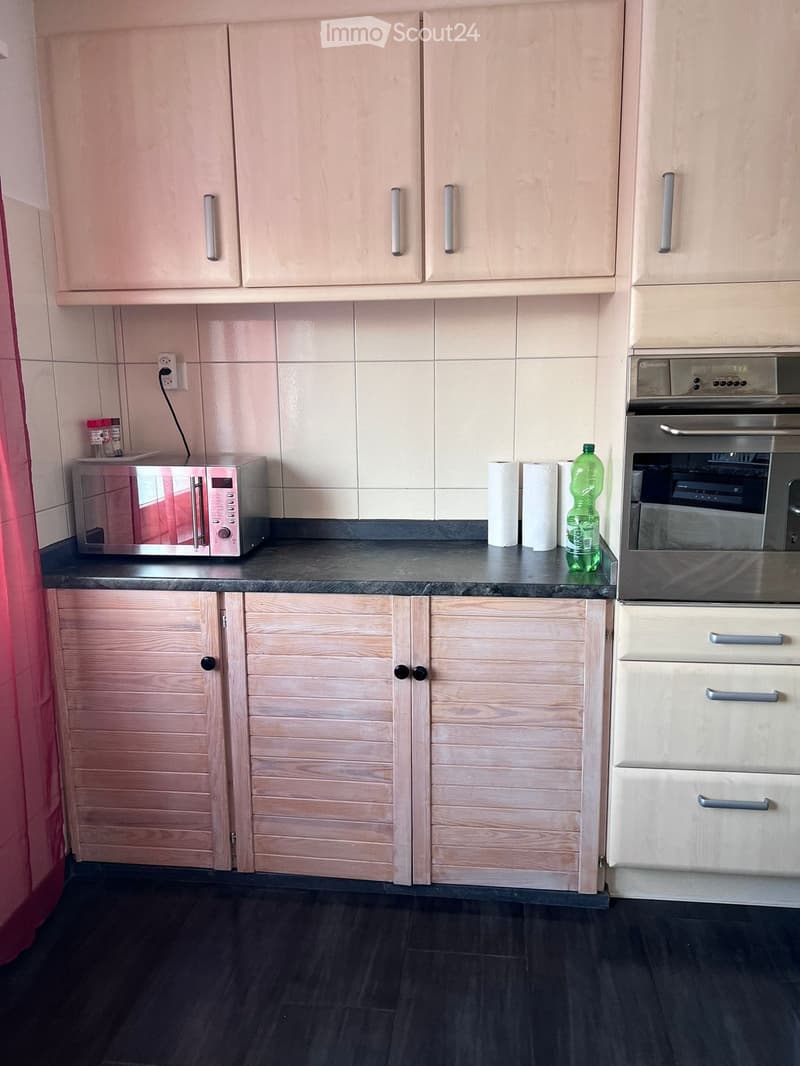 Modern kitchen with light wood cabinets, black countertops, and stainless steel appliances including a microwave and oven. A bottle and paper towels are on the counter.