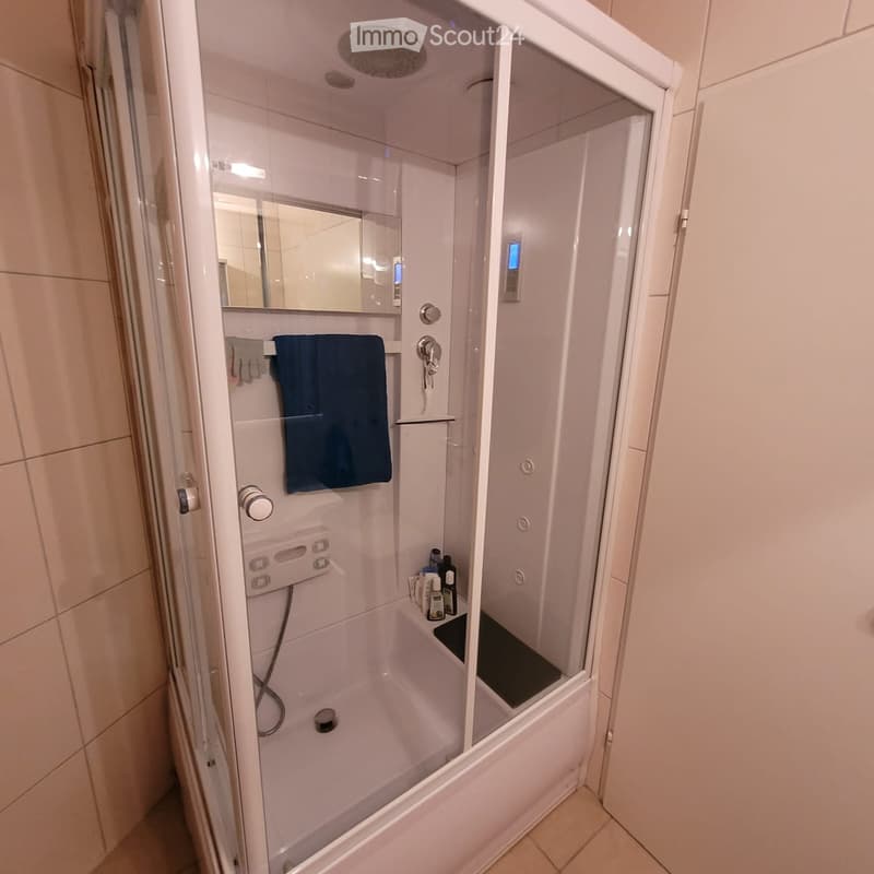 Modern walk-in shower with glass enclosure, built-in seat, and control panel. Features a towel rack, mirror, and multiple shower heads.