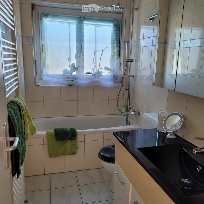 Modern bathroom with a bathtub, toilet, sink, and a mirror. Green towels are placed on the edge of the bathtub and a window with blinds allows natural light to enter.