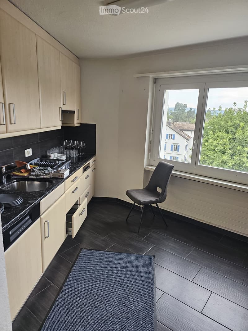 Modern kitchen with light wood cabinets, black countertops, and stainless steel appliances. Features a sink, faucet, dish rack, wine glasses, a chair, and a window with a scenic view.