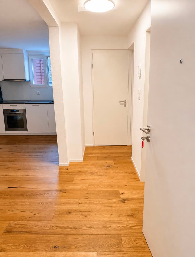 Empty hallway with wooden flooring, white walls, and a closed door at the end. A kitchen area is visible to the left with white cabinets and a built-in oven.