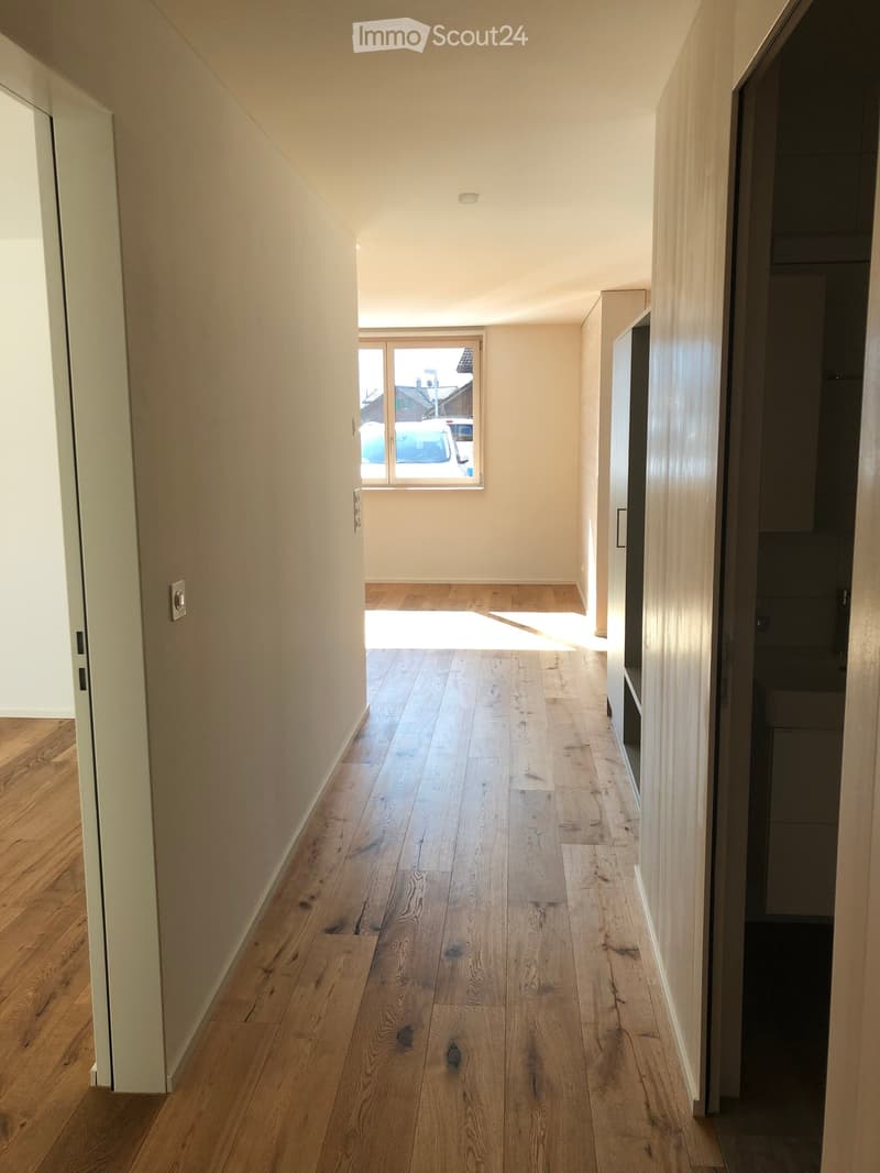 Empty hallway with light wooden flooring, white walls, and a window at the end letting in natural light.