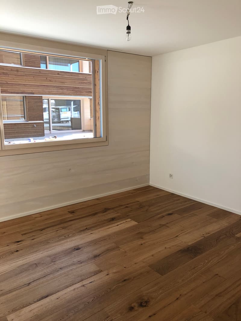 Empty room with wooden flooring, white walls, and a large window with wooden frame showing an outdoor view.