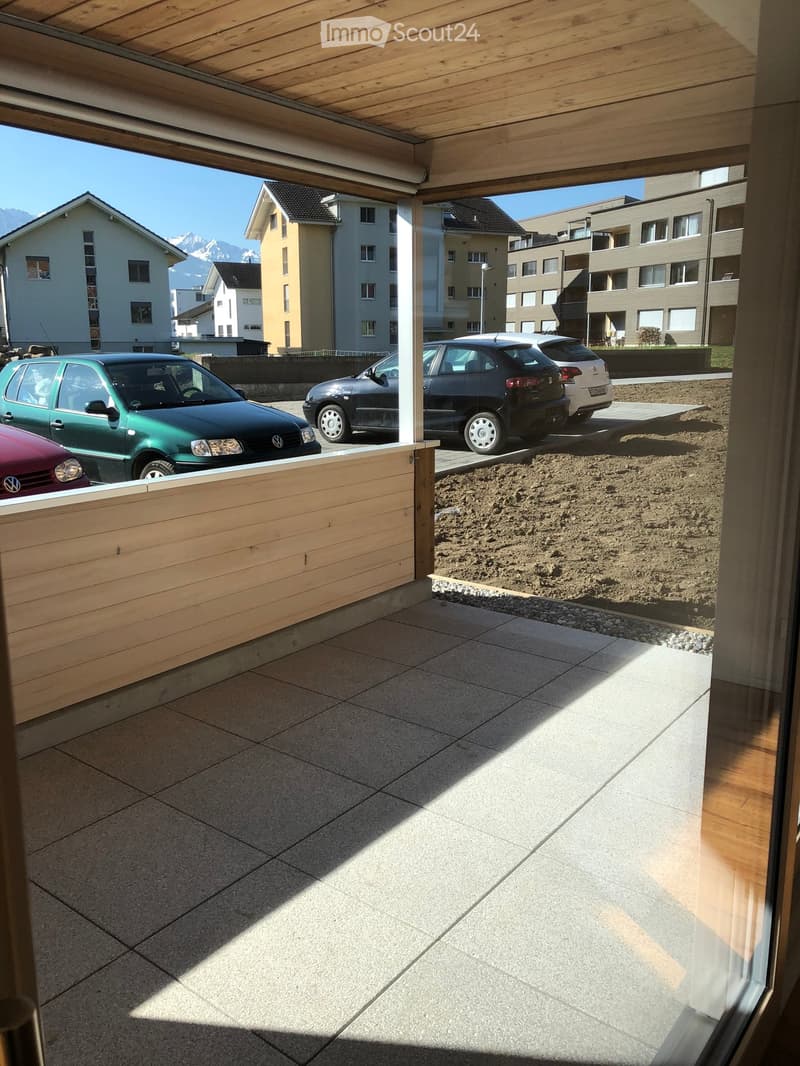 Balcony with wooden decking, tiled floor, and wooden railing, overlooking a parking area with several cars and surrounding buildings.