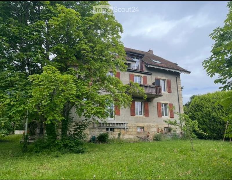 A two-story house with red shutters and a stone facade, surrounded by lush greenery and trees.
