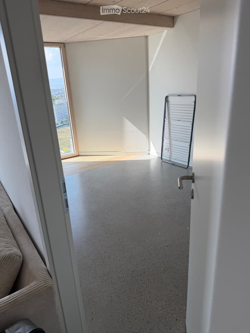 Empty room with concrete floor, white walls, large window with view, and a clothes drying rack in the corner.