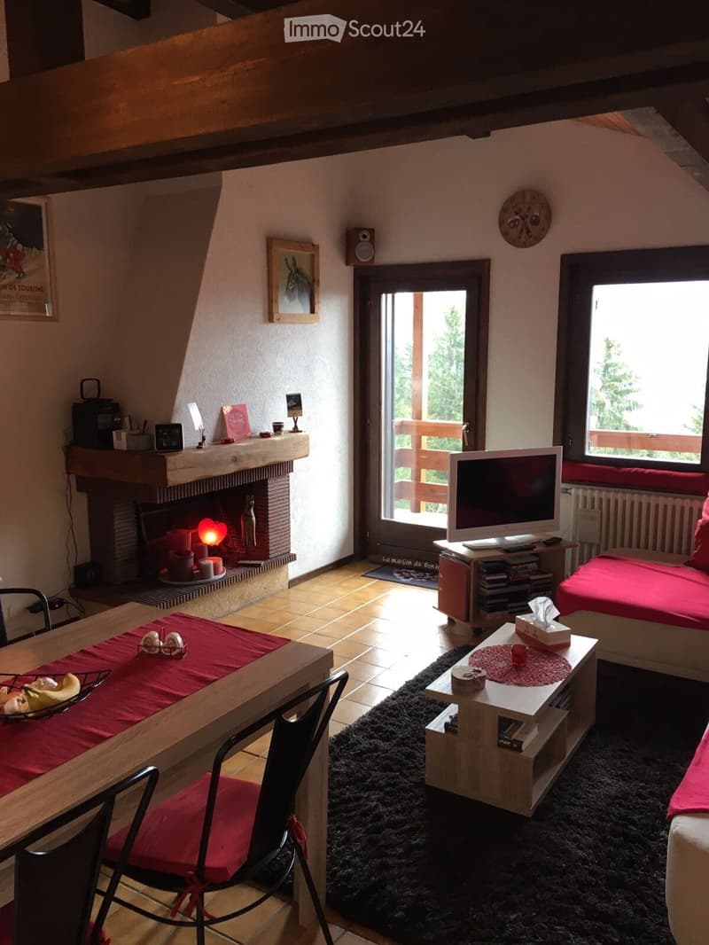 Cozy living room with a fireplace, sofa, dining table, and television. The room features wooden beams, large windows, and a balcony door leading outside.