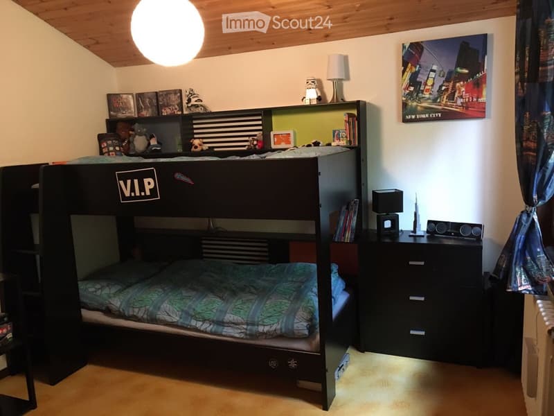 A modern bedroom with a lofted twin-sized bed, nightstand, bookshelf, and decorative items. The room features a large window with curtains and a poster on the wall.