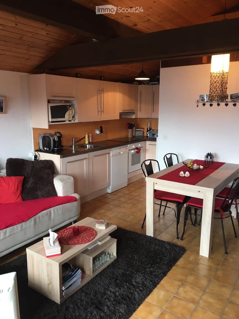 A modern, open-plan living and dining area with a kitchen. Features include a white sofa with red cushions, a wooden coffee table, a dining table with chairs, and a fully equipped kitchen with white cabinets, a microwave, dishwasher, and oven. The space h