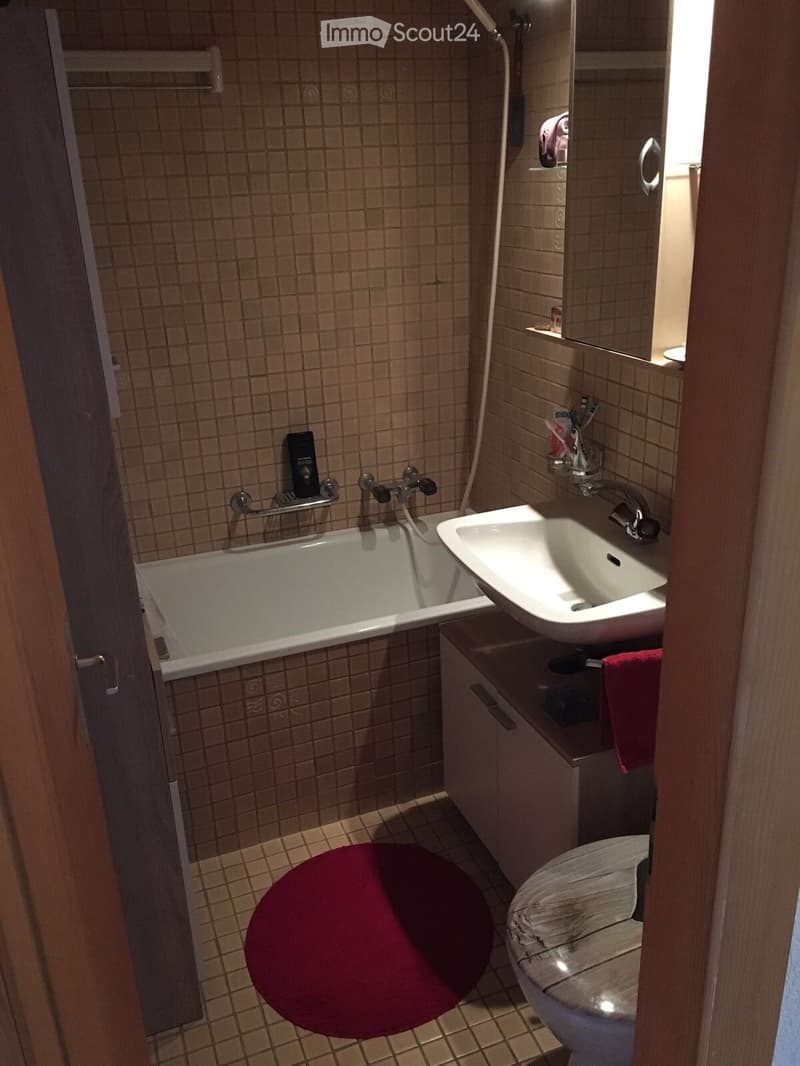 Bathroom with a bathtub, sink, toilet, and mirror. Features tiled walls and floor, a wooden cabinet, and a red towel on the floor.