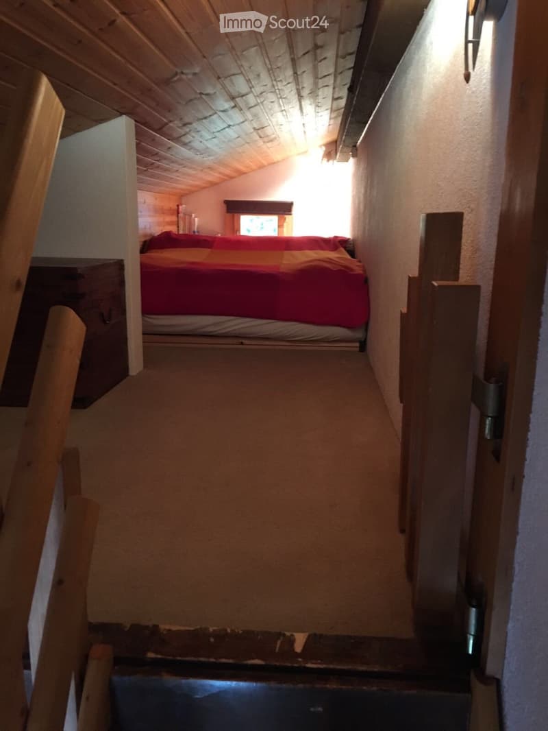 Cozy attic bedroom with wooden beams, a single bed with red and yellow bedding, and a small window letting in natural light.