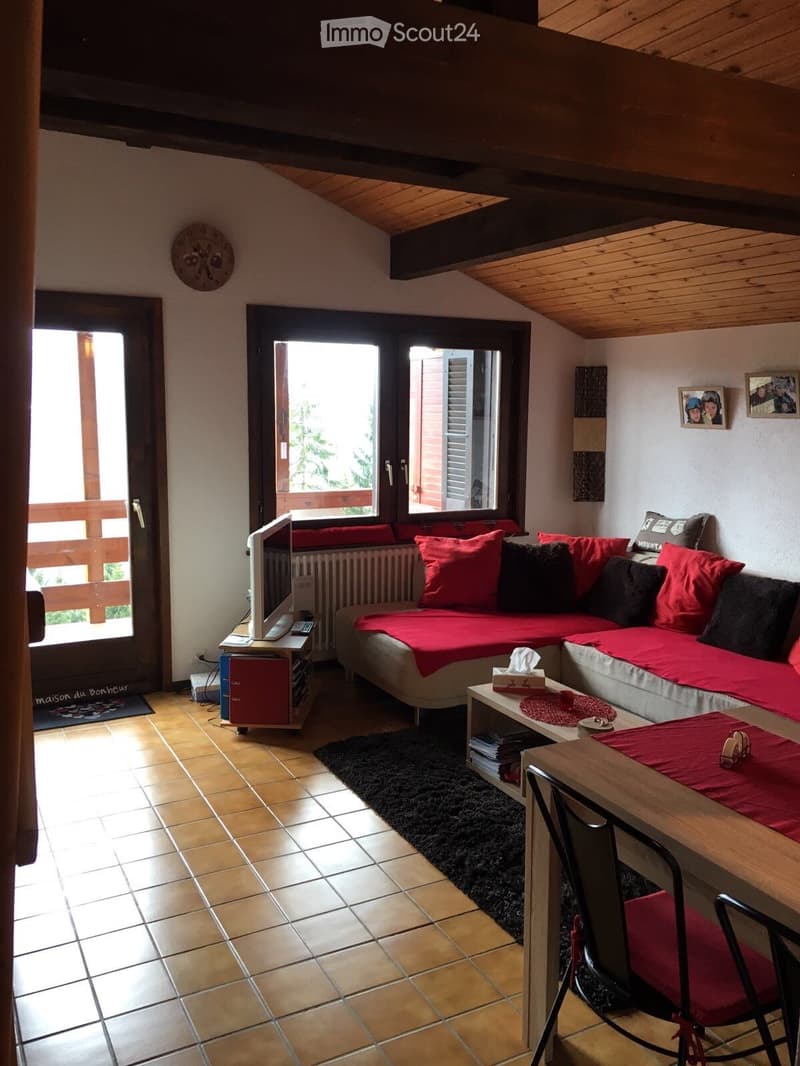 Cozy living room with a sectional sofa, coffee table, TV, and a dining area with a table and chairs. The room features wooden beams on the ceiling and large windows with a balcony view.