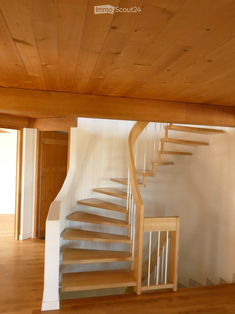 Spiral staircase with wooden steps and a white railing, leading upwards in a well-lit hallway with wooden flooring and white walls.