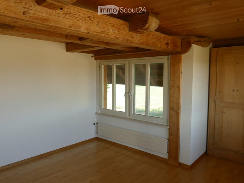 Empty room with wooden floor, white walls, exposed wooden beams on the ceiling, and a large window with multiple panes.
