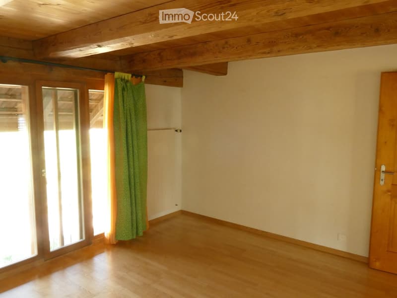 Empty room with wooden flooring, exposed wooden beams on the ceiling, and a large window with green curtains partially drawn.