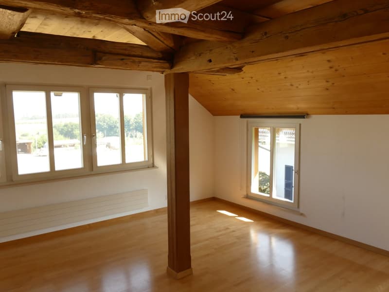 Empty room with wooden floor, exposed wooden ceiling beams, large windows, and a central support column.