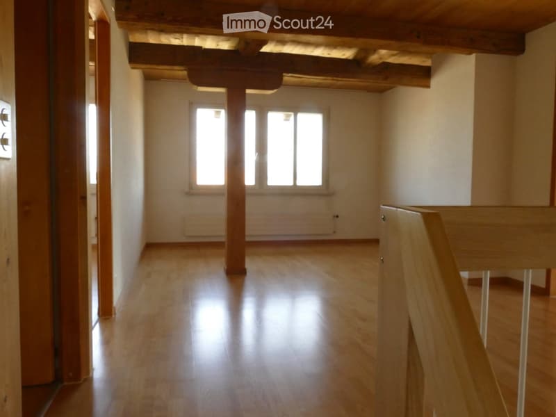 Empty room with wooden flooring, exposed wooden beams on the ceiling, and a large window allowing natural light to enter.
