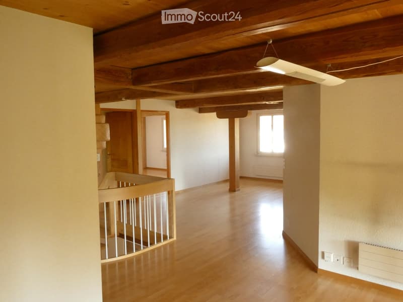 Empty room with wooden floor, exposed wooden beams on the ceiling, white walls, and a staircase leading to another level.
