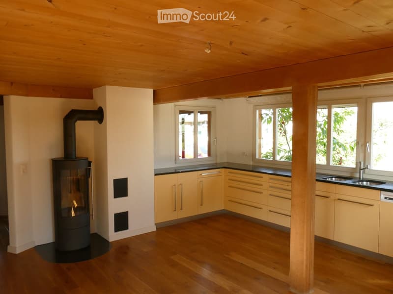 Modern kitchen with light wooden cabinets, a black countertop, and a wood-burning stove. The room features a wooden ceiling and floor, with large windows allowing natural light to fill the space.