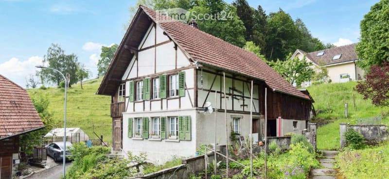 Traditional Swiss house with a half-timbered facade, green shutters, and a red-tiled roof, surrounded by greenery and a small garden.