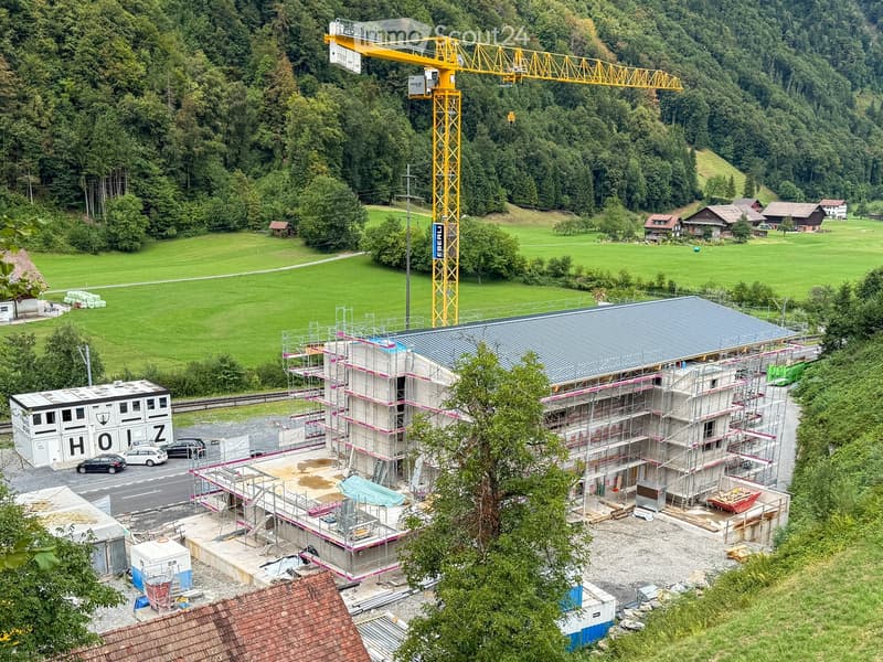 A multi-story building under construction with scaffolding, surrounded by greenery and a crane in the background.