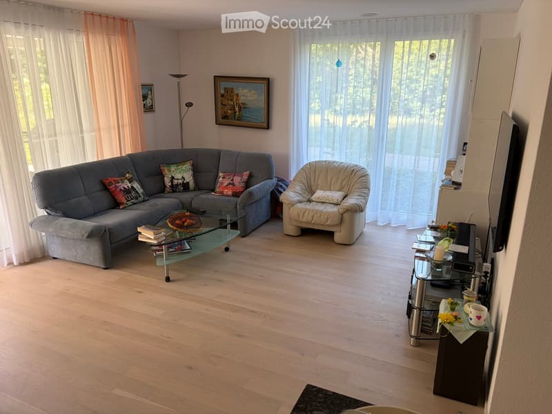 Bright living room with wooden flooring, large windows, a grey sectional sofa, a beige armchair, a glass coffee table, a TV stand, and decorative items including books, a flower vase, and picture frames.
