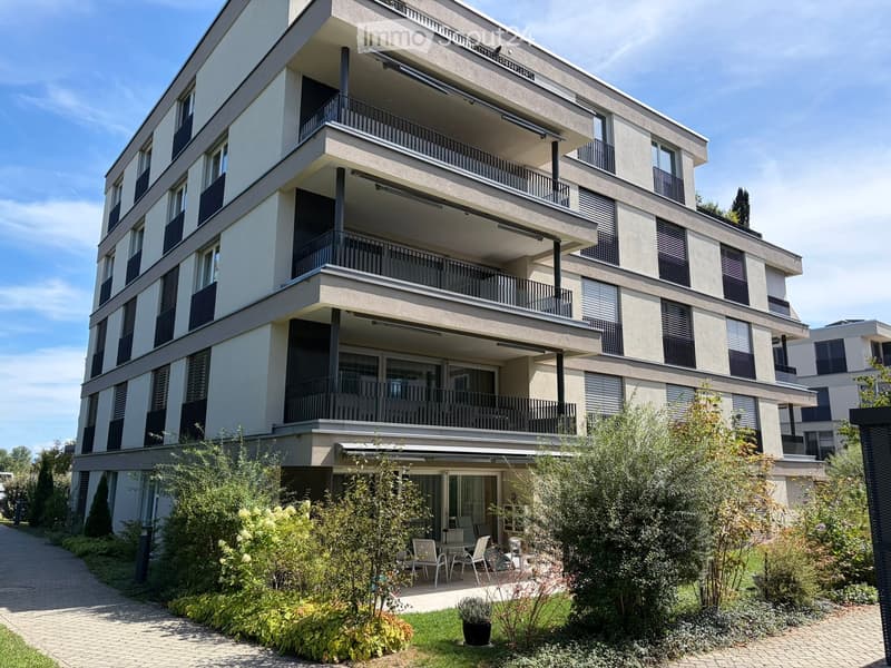 Modern multi-story residential building with balconies, large windows, and surrounding greenery including chairs and tables in the courtyard.