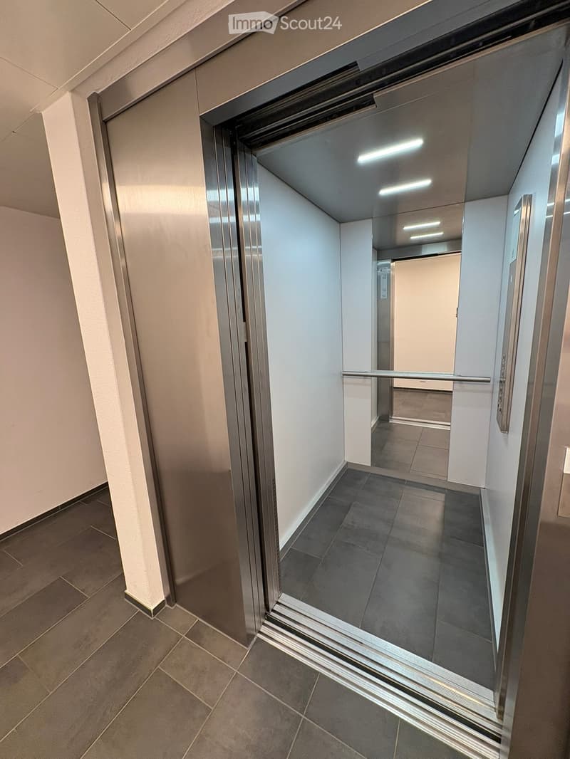 Modern hallway with tiled floors, white walls, and an elevator with glass doors. The hallway is clean and well-lit with recessed lighting.