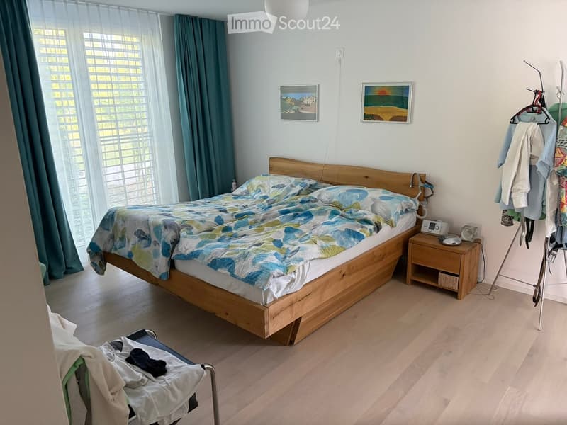 Bright bedroom with wooden flooring, large window with blinds, bed with colorful bedding, nightstand with lamp, clothing rack, and picture frames on the wall.