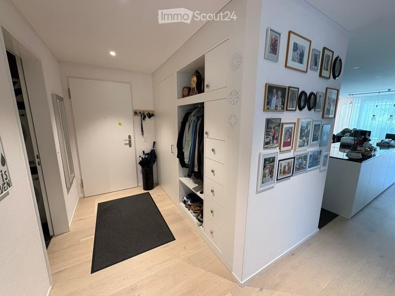 Modern hallway with light wooden flooring, white walls, built-in closet with shoes and coats, framed photos on the wall, and a small black rug near the entrance.