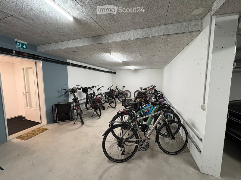 Underground parking area with multiple bicycles parked in rows, concrete floor, white walls, and a door leading to another area.