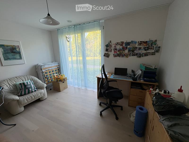 A multi-functional room with a sofa, desk, laptop, chair, and various personal items. The room features large windows with curtains, a picture frame on the wall, and a yoga mat on the floor.