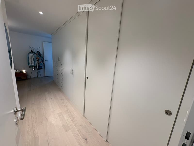 A clean, modern hallway with light wooden flooring, white walls, built-in white closet units with multiple drawers, and a small side table with a lamp and some items on it.